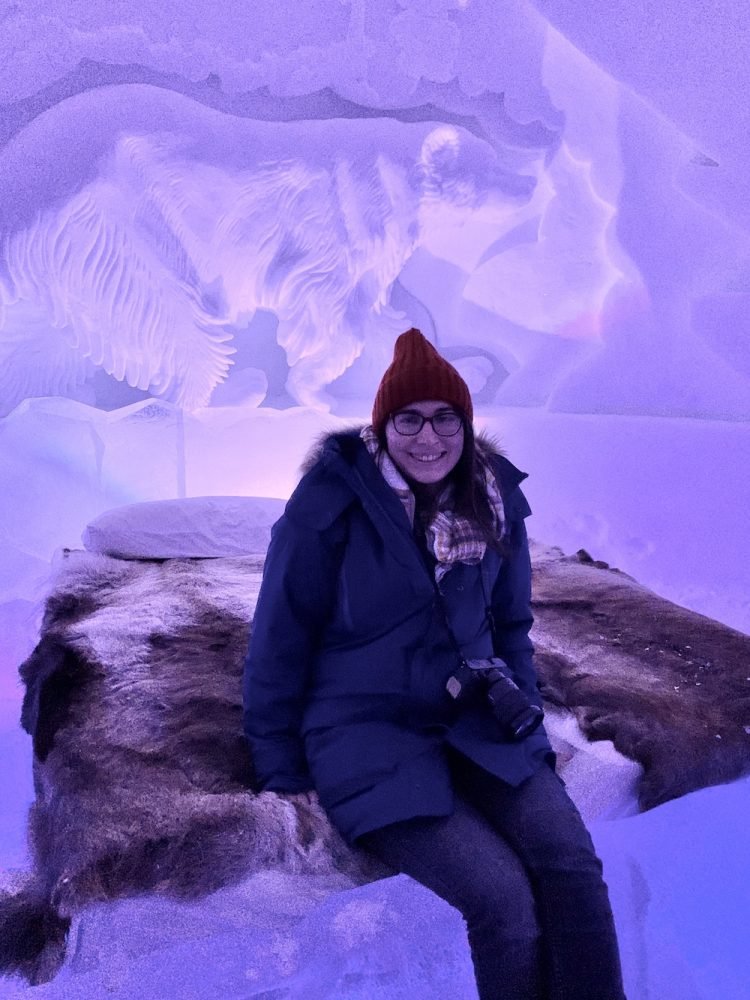 Allison Green sitting on an ice bed covered in a reindeer pelt at a ice hotel, wearing a red hat, scarf, parka, and a large camera. She is smiling and there are intricate ice sculptures in the background.