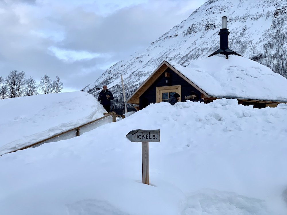 Entrance to the Tromso Ice Domes tickets center, with sides of the pathway totally covered in snow, and a small sign that says 'tickets' leading the way.