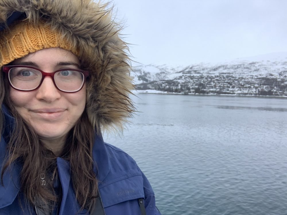 Allison Green on a fjord cruise in Tromso during the winter, wearing a yellow hat, parka with faux fur, and red glasses.