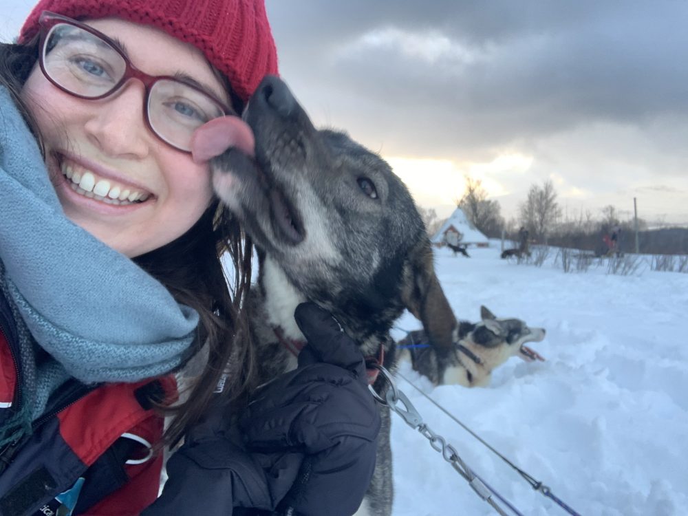 Allison Green in Tromso, Norway, with a happy husky licking her face after doing a dog sledding tour. She is wearing a red hat and blue scarf and a big smile.