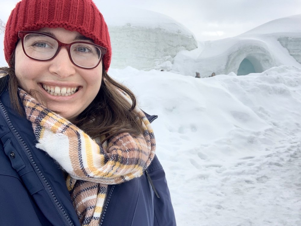 Allison Green wearing a red beanie, plaid scarf, blue jacket and smiling at the camera while visiting the Tromso Ice Domes in Northern Norway, a famous ice hotel