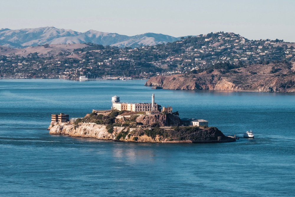 A small island, Alcatraz, with an abandoned prison building on it, in the middle of beautiful turquoise water surrounded by a city.