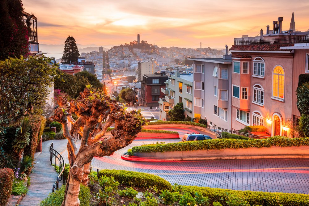 A view of the crooked turns of Lombard Street, lit up at night with a light trail from a car, with a view of Coit Tower off in the distance.