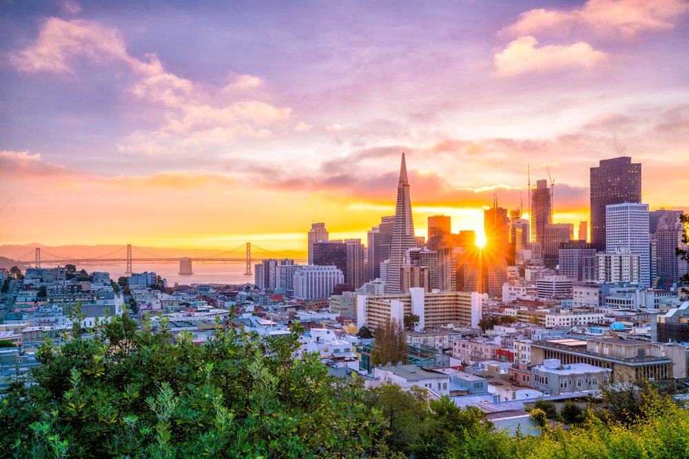 A view of the San Francisco skyline from up on a hill with greenery in the foreground and a sunburst seen through the skyline at sunset, a view of a bridge in the distance.