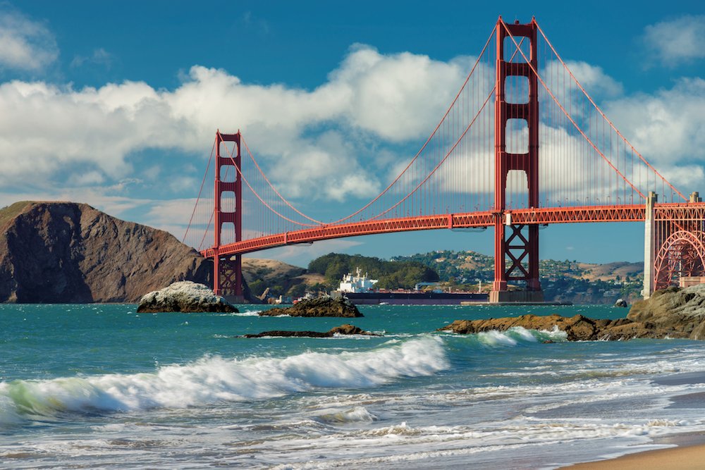 View of waves crashing on the beach with a view of the red suspension bridge, the Golden Gate Bridge, over turquoise water on a partly cloudy day.