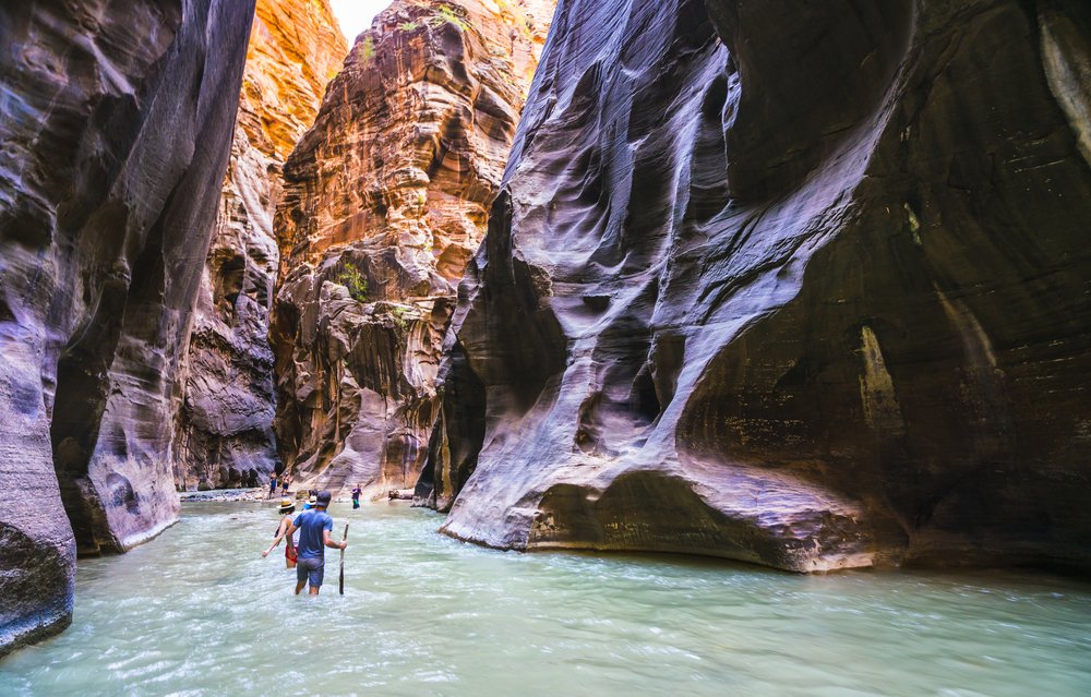 People hiking in knee-deep water in hiking sticks in a slot canyon with purplish rocks and pale green water.
