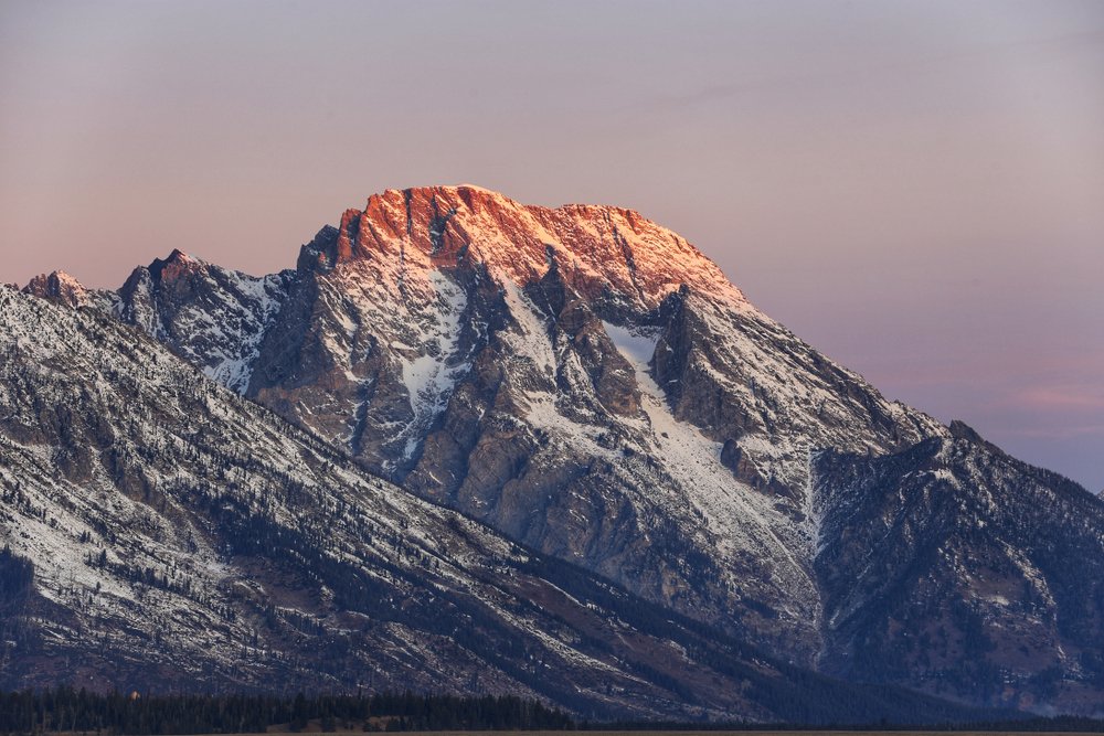 Alpenglow (the reddish glow at sunrise on mountain peaks) illuminating a peak of the Teton Range with a pastel lavender sky.