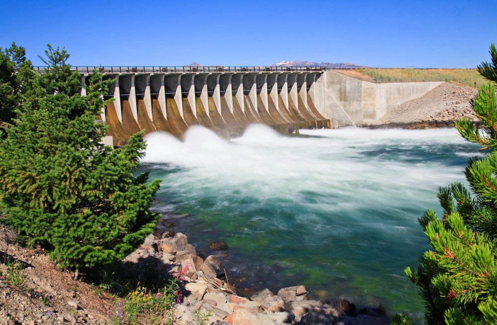 A very large cement dam showing rushing water in a long exposure photo rushing underneath the dam, turquoise water coming from the dam, and pine trees on the sides of the dam.