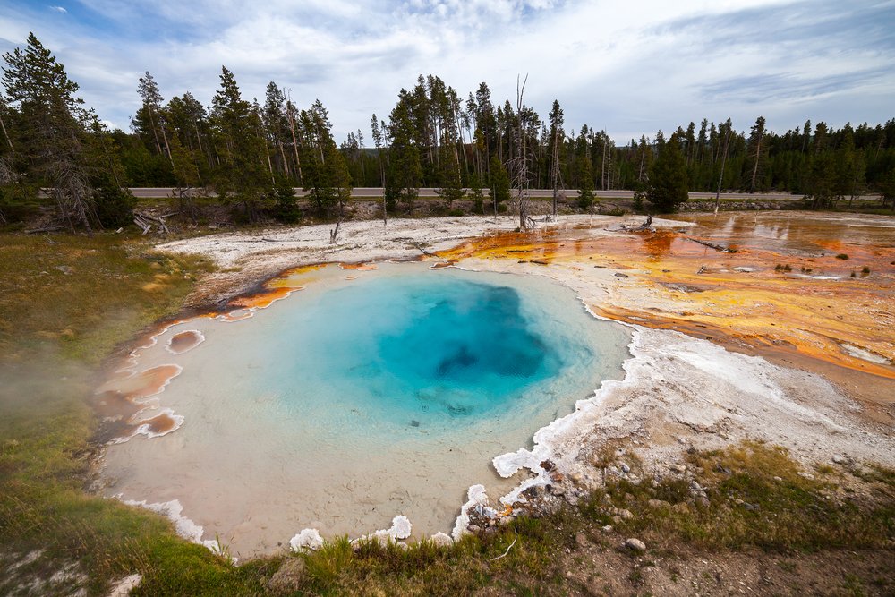 A geyser in Yellowstone, orangeish deposits on one side of it with a deep blue spring in the middle.