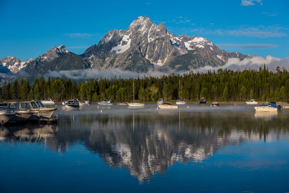 A mountain perfectly reflecting in the still water at Colter Bay, with lots of boats sitting still in the water, anchored.