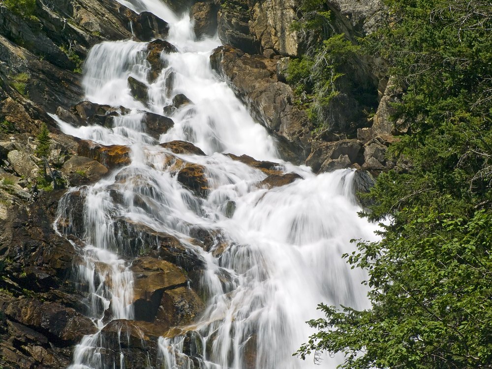 Close up of a section of a waterfall cascading down rocks with some green trees in the foreground.