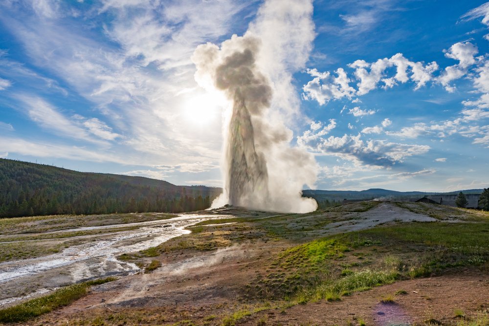 A geyser of steam bursting a hundred feet into the air, surrounded by a barren landscape, on a partly cloudy day with afternoon light.
