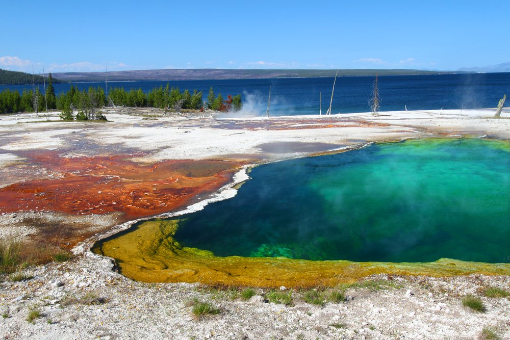 A deep blue and turquoise geyster, with orange and white mineral deposits beside it, next to a deep blue lake.