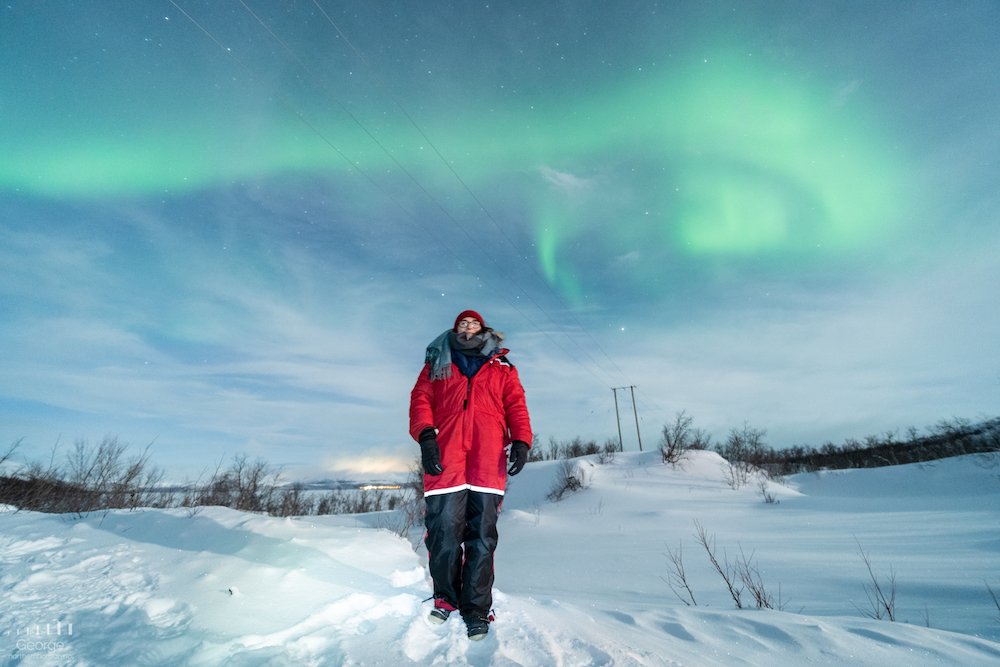 Allison Green posing with the Northern lights on a tour in Norway in a red thermal suit with the green aurora overhead, surrounded by snowfall