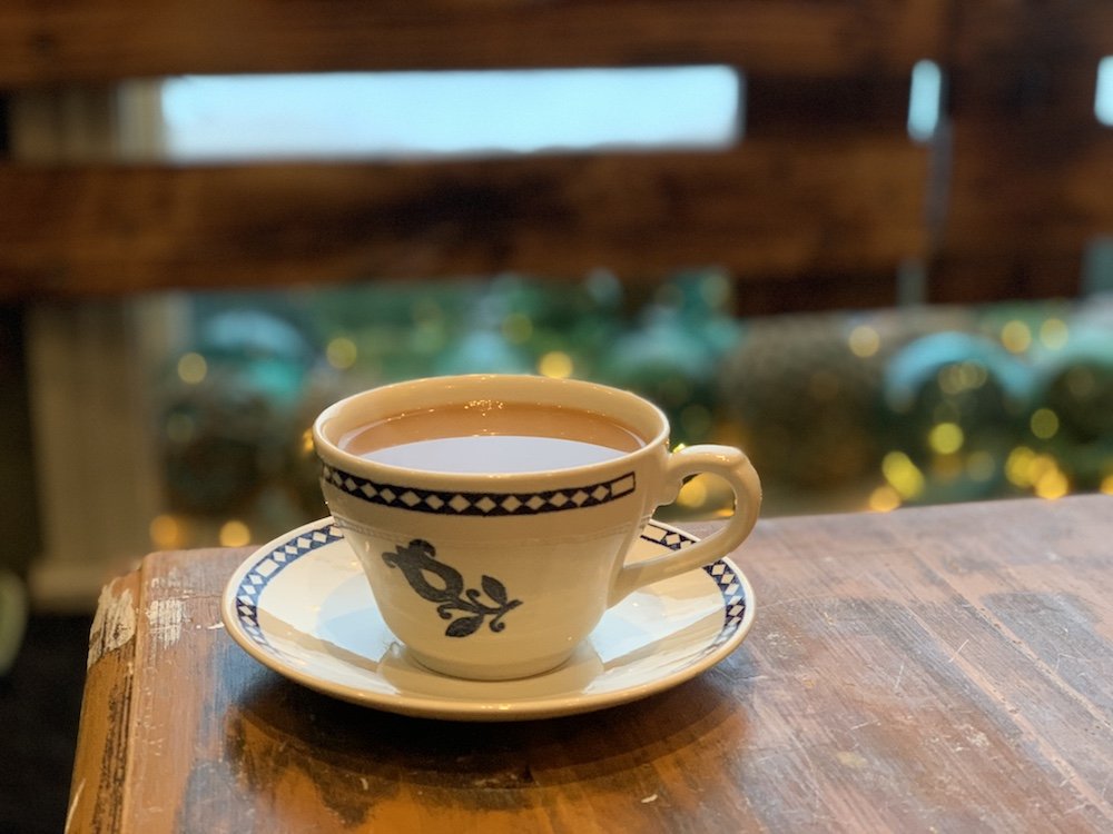 A cup of coffee served in a tea-cup style cup with a saucer, with festive decorations behind.