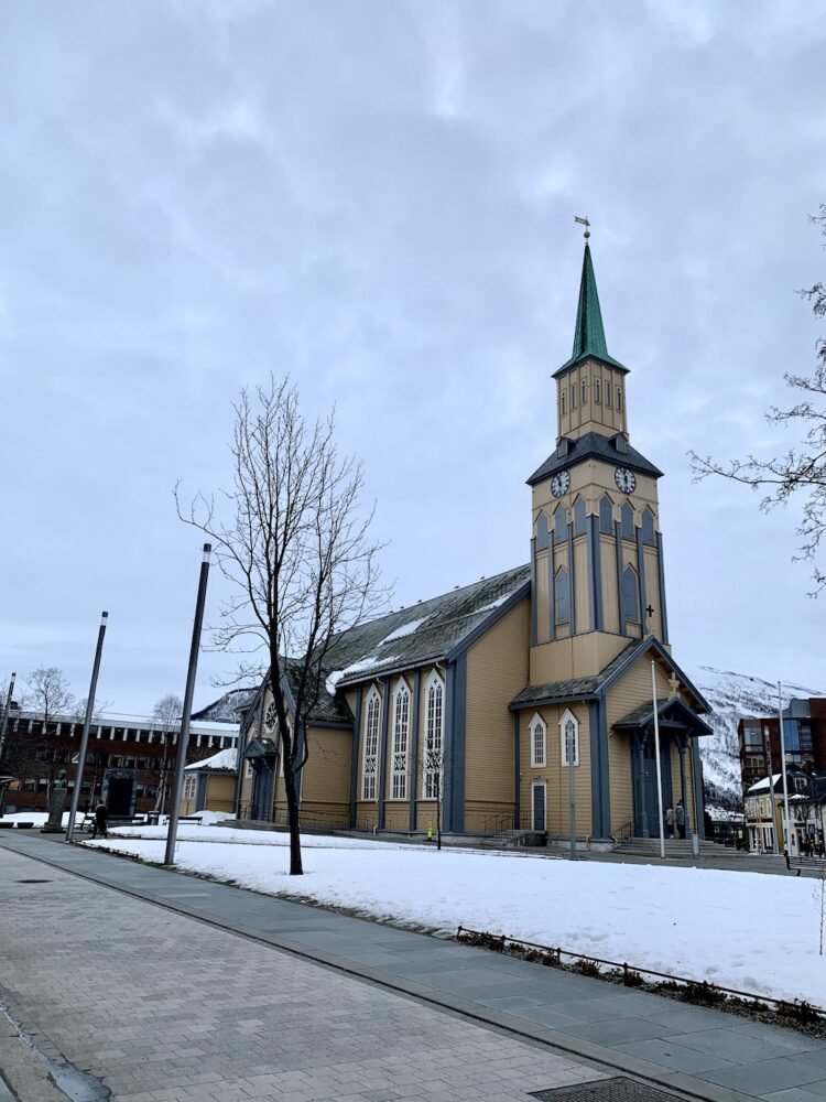 the winter light falls on the beige and brown tromso cathedral with a clocktower and single spire in the downtown area, where the sidewalks are clear of snow but the park has some snowpack