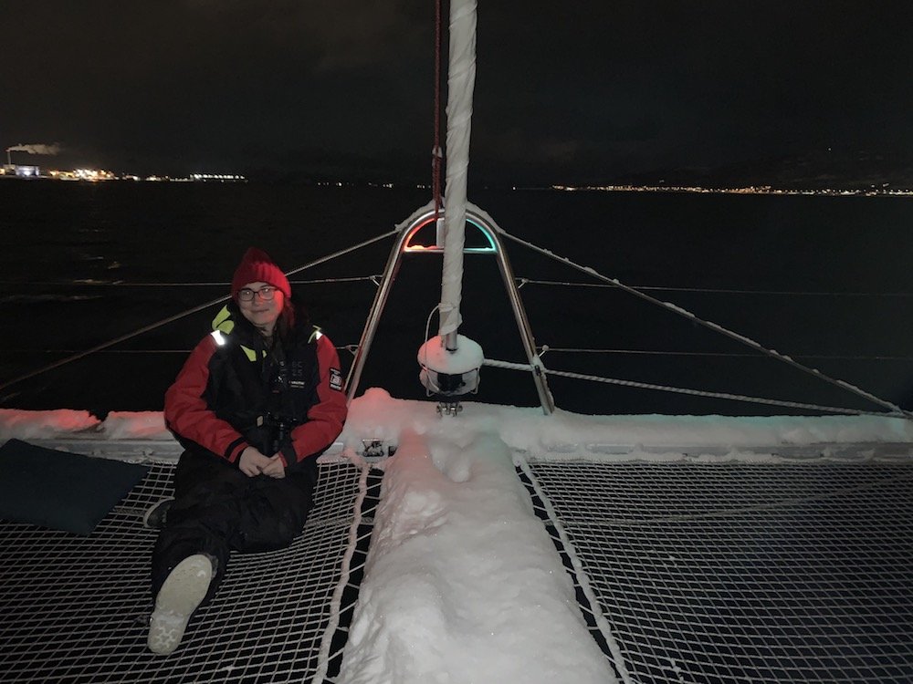 Allison Green sitting in a polar thermal suit, with a hat on, on a catamaran net on a snow-covered boat