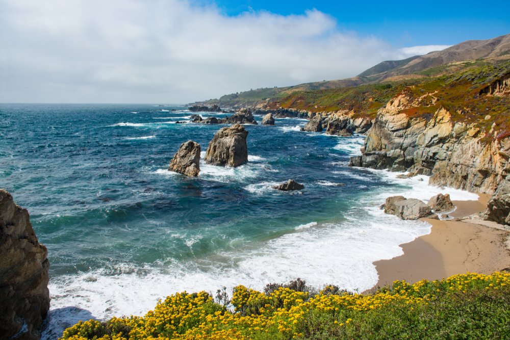 the sandy shore and blue ocean waters of the rugged garrapata beach with rock outcroppings in the water