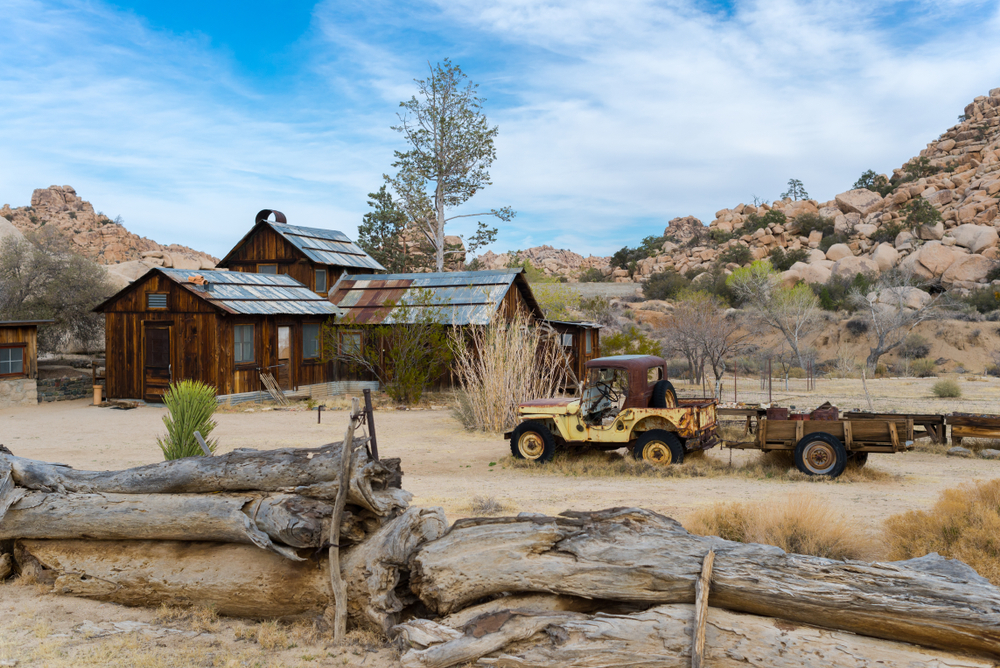 A broken down car and the remnants of a ranch (wooden house with tin roof) in the middle of Joshua Tree's desert landscape on a partly cloudy day.