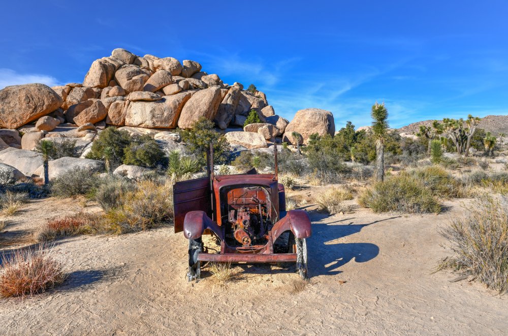 A broken down, rusted over red car in the middle of a Joshua Tree landscape.