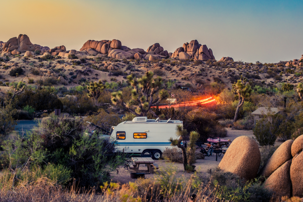 An occupied RV with the lights on in the landscape of Joshua National Park with some light trails in the background from a moving car near sunset.