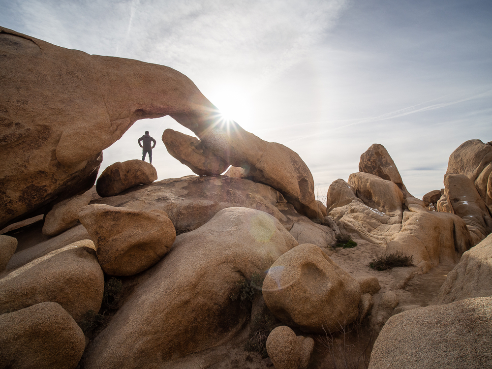 A man standing underneath a rock arch with a foreground of lots of smooth rocky boulders on a partly cloudy day, with a sunburst hitting the rock arch.