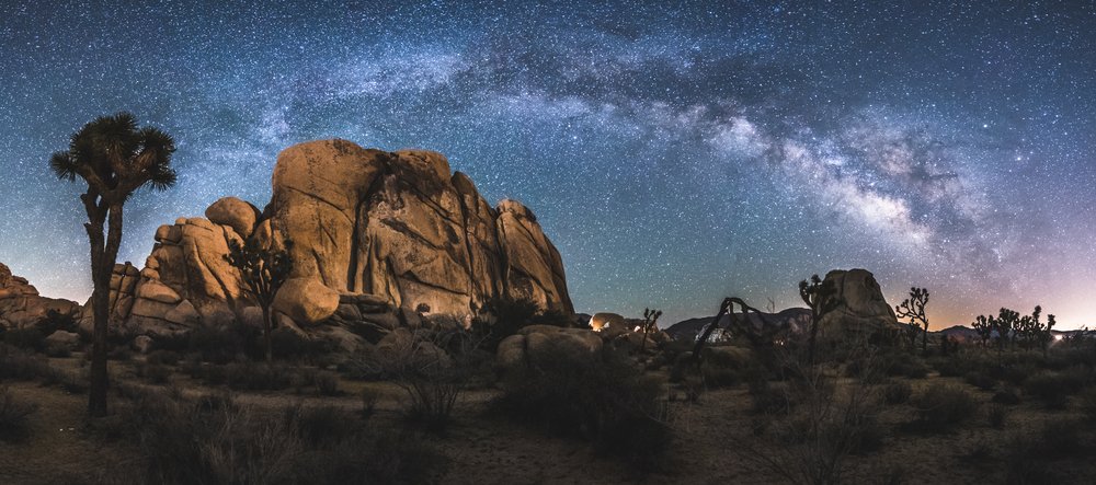 A panoramic view of the rocks and trees of Joshua Tree National Park with the milky way lit up overhead in colors of lavender, pink, and blue.