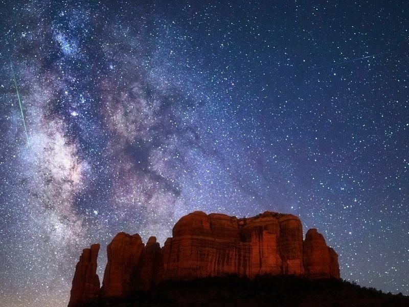 The Milky Way shown in the night sky above the red rocks of Sedona's landscape with light trails from a plane or shooting star