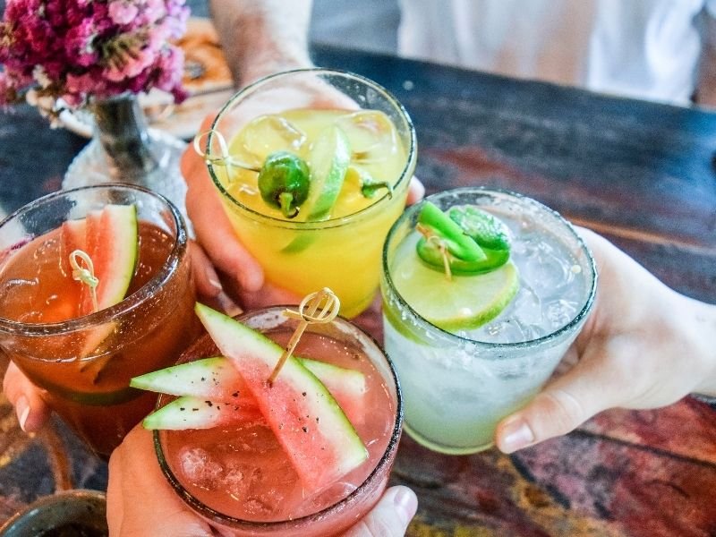 Four hands holding colorful drinks including one with watermelon and one with lime.