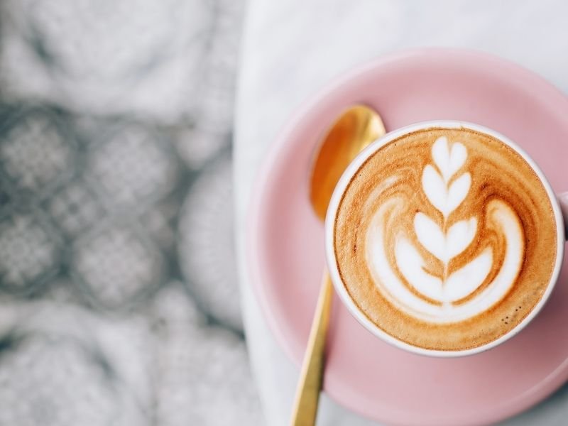 Perfect latte art in a cup on a pink plate with a gold spoon on a table.