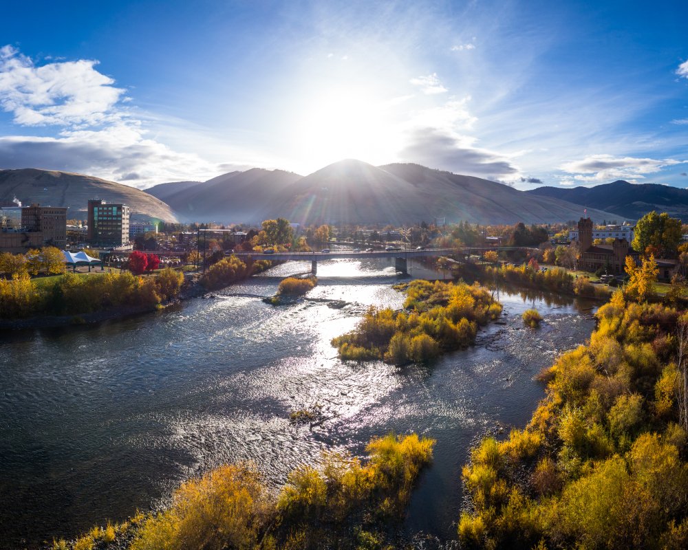 A sunny view of the sun setting below Missoula mountain ranges with some buildings on one side, the river in the middle, a bridge crossing the river, and trees on the other side of the river.