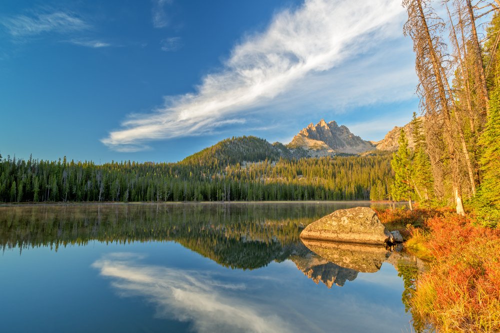 A lake with a perfect reflection on the evergreen trees and mountains in the still water, some yellow and orange fall foliage in the left corner.