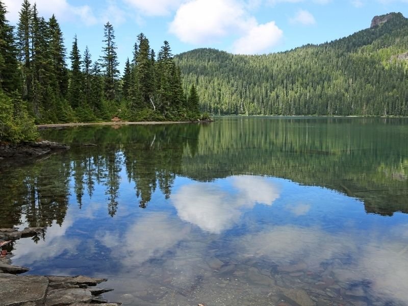 A calm lake reflecting mountains and clouds and trees in Mt. Rainier National Park, called Mowich Lake.