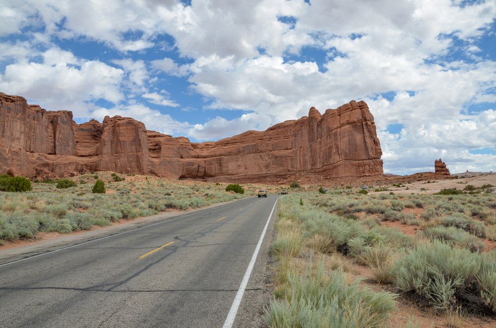 large red sandstone 'wall' next to a road with a car on it driving in arches national park on a sunny partly cloudy day.