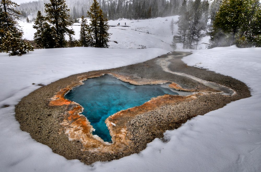 Snow covered landscape revealing a geyser in colors of orange on the rim and deep turquoise blue in the middle. Pine trees in background.