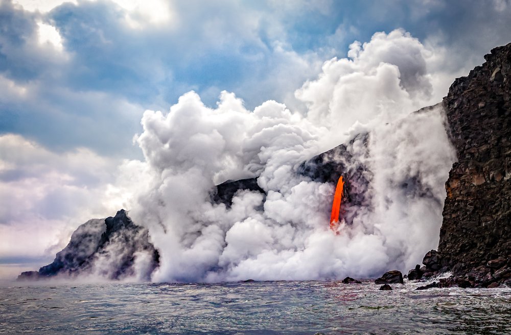 Cloudy sky with a view of orange lava flowing into the Pacific Ocean, causing a large cloud of white-gray smoke at the place where lava meets water.