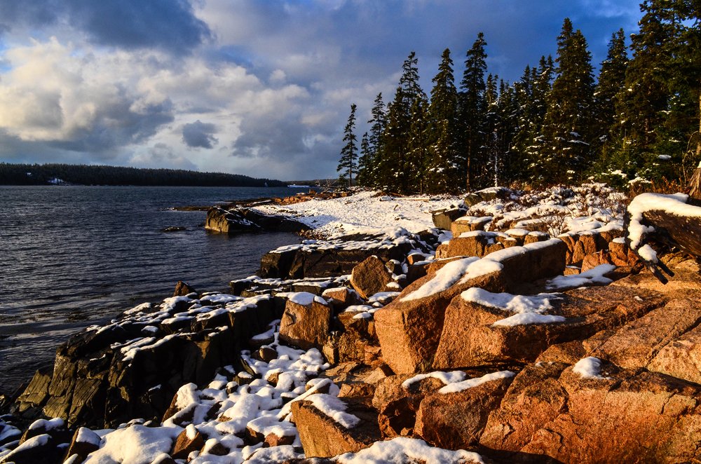 Snow covered rocks with background of Atlantic Ocean and trees.