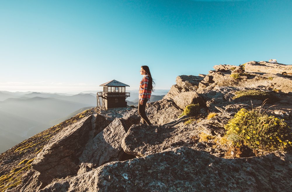 A woman wearing a plaid shirt and black pants looking off into the distance of the mountains with a fire lookout (a small wooden hut) in the background.