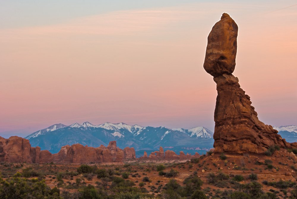 hoodoo holding up a rock that looks like its balancing. mountains capped with snow in the distance at sunset.