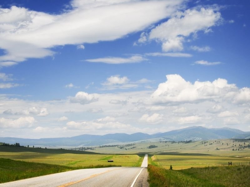 A rolling highway road leading towards Big Sky Montana mountain resort, grassy fields leading to mountains in the distance.