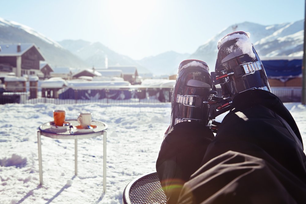 Feet up with ski shoes still on with apres ski drinks in the background and a view of distant ski chalets and mountains