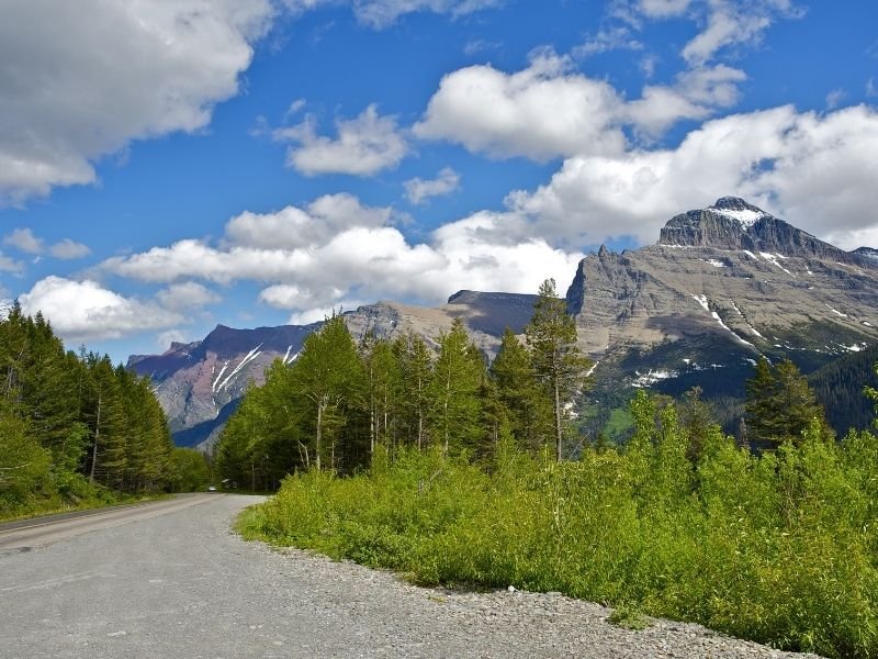 A road near Glacier National Park with greenery and some mountains with a bit of snow nearby