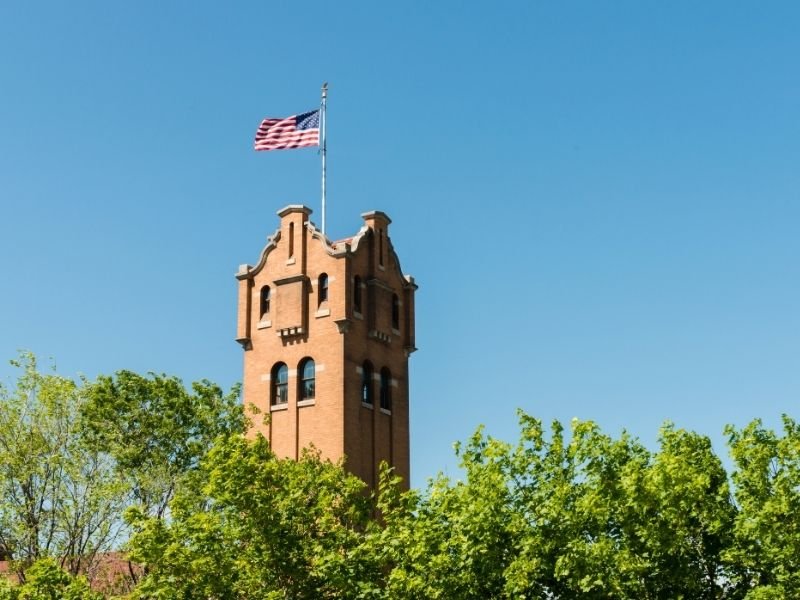 A building tower with an American flag on it raised above the tops of green trees on a blue sky cloudless day.