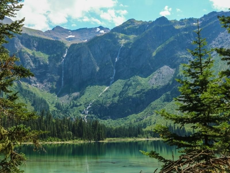 A deep teal and turquoise glacial lake, surrounded by pine trees and steep mountains with some waterfalls coming down the sides from snow melt.