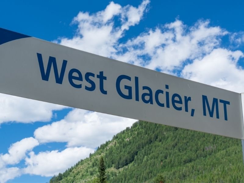 Sign which reads "West Glacier, MT" against a backdrop of a partly cloudy sky and green trees.