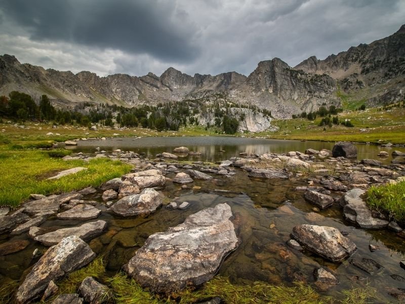 Lots of rocks in a shallow water pool surrounded by rocky mountains and green grass on an overcast day hiking