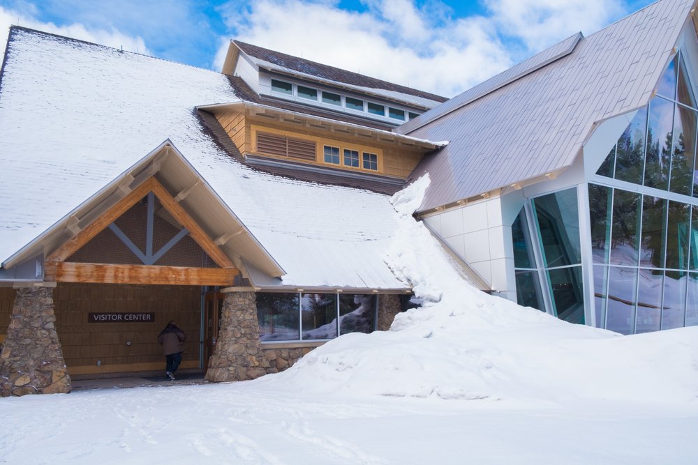 Wood, stone, and glass building with snow piled high and on roof with the words "Visitor Center" and one person entering the building