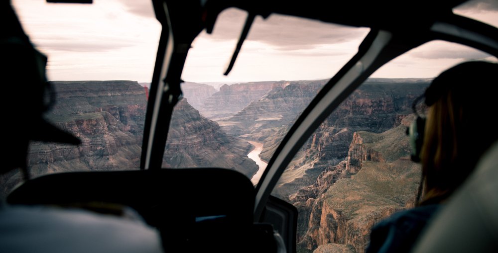 View from helicopter cockpit of man and woman in helicopter wearing headphones with Grand Canyon in background.