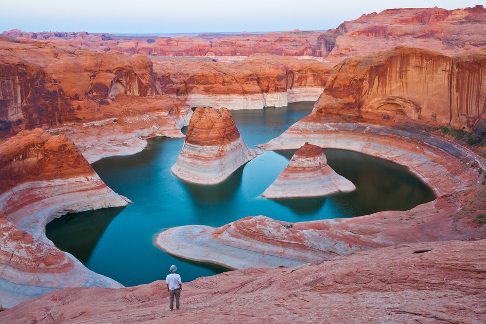 A man standing with a view of Glen Canyon's glassy still water and red rock landscape in front of him at Reflection Canyon.