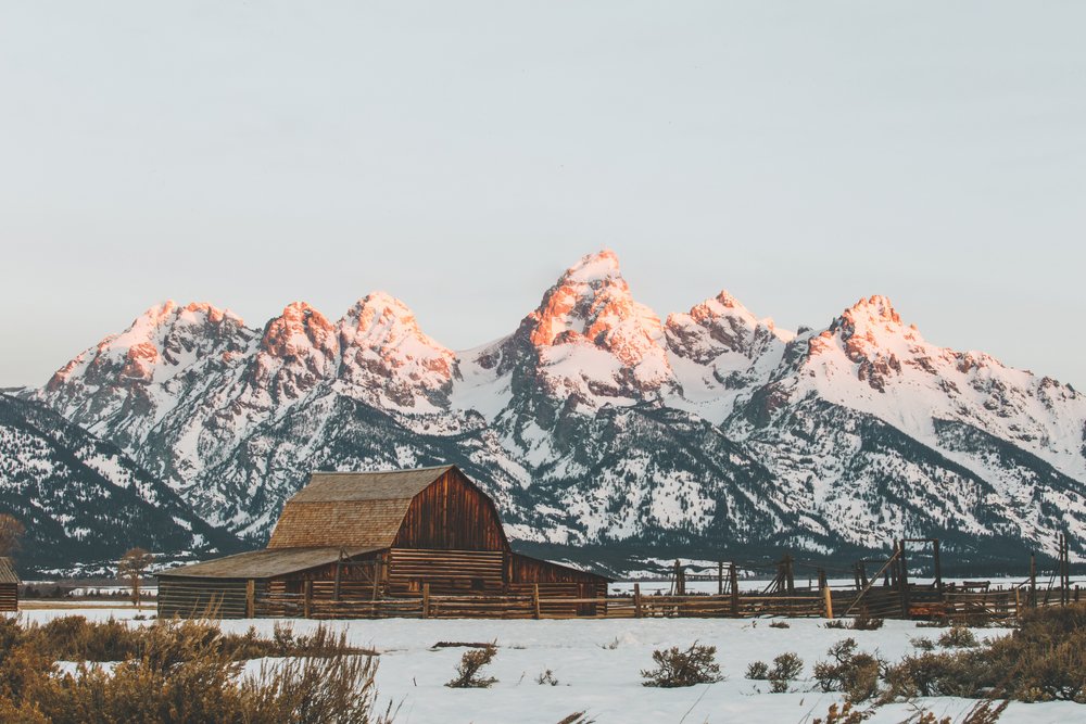 A view of the famous "Grand Teton Cabin", a wooden structure shaped almost like a sombrero hat, with a sunrise light glow on the tips of the mountain range behind.